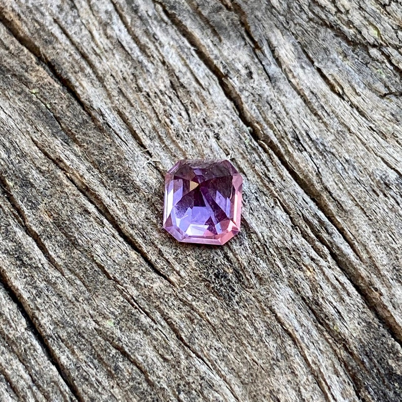 Puede incluir: Una piedra preciosa facetada de forma cuadrada con un tono p&uacute;rpura y rosa. La piedra preciosa est&aacute; colocada sobre un fondo de madera.