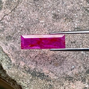 May include: A vibrant pink rectangular gemstone with a slightly cloudy appearance, held in a pair of tweezers.