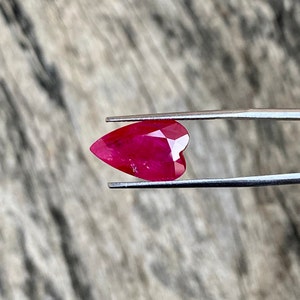 May include: A heart-shaped, faceted ruby gemstone held by tweezers. The ruby is a deep, vibrant red color with multiple facets that catch the light. The tweezers are silver-colored and hold the gemstone against a neutral background.