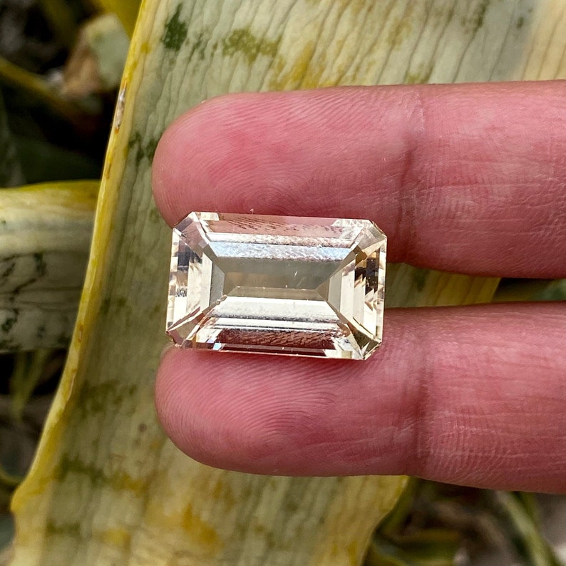 May include: A clear, rectangular, faceted gemstone with a light peach hue. The gemstone is cut in an emerald shape.