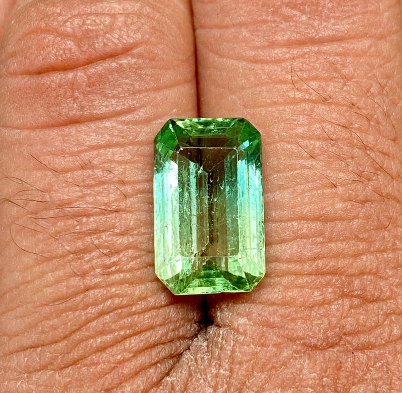 May include: A vibrant green emerald gemstone cut in an emerald shape. The gemstone is approximately 10mm x 7mm and has a brilliant, faceted finish.