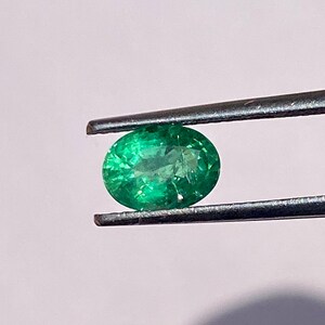 May include: A vibrant green emerald gemstone held by tweezers. The emerald is round and has a smooth, polished surface.