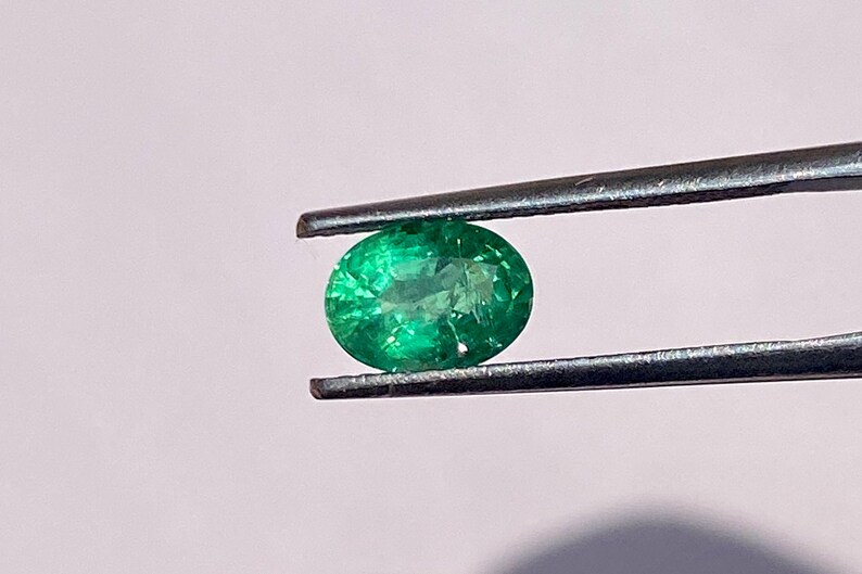 May include: A vibrant green emerald gemstone held by tweezers. The emerald is round and has a smooth, polished surface.