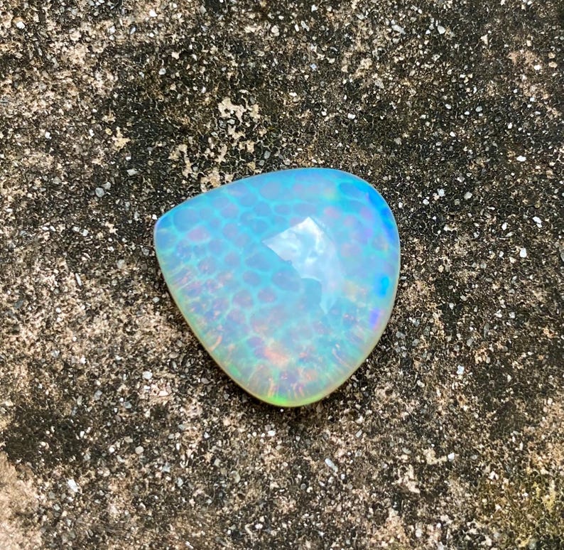 May include: A light blue iridescent opal gemstone with a teardrop shape. The opal has a white base with a blue and green iridescent sheen.