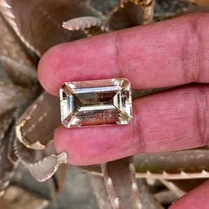 May include: A clear, rectangular, faceted gemstone with a light yellow hue. The gemstone is held in a hand.