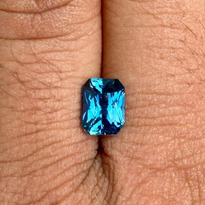 May include: A vibrant blue gemstone with a rectangular emerald cut. The gemstone is set in a simple, clear setting and is displayed on a finger.