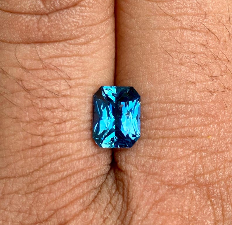 May include: A vibrant blue gemstone with a rectangular emerald cut. The gemstone is set in a simple, clear setting and is displayed on a finger.