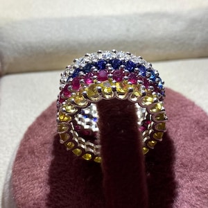 Könnte beinhalten: Ein mehrfarbiger Edelsteinring mit einer Reihe blauer Saphire, einer Reihe roter Rubine und einer Reihe gelber Saphire. Der Ring ist in Weißgold gefasst und hat eine polierte Oberfläche.