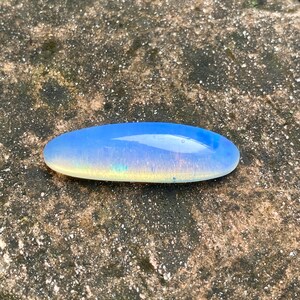 Puede incluir: Una piedra preciosa de ópalo ovalada lisa, de color azul claro con colores arcoíris iridiscentes.