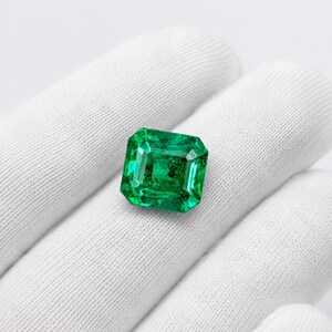 May include: A vibrant, emerald-green gemstone cut into a rectangular shape with faceted edges. The stone displays a deep, saturated green hue with internal clarity and light-reflecting surfaces, showcasing a classic step-cut design.
