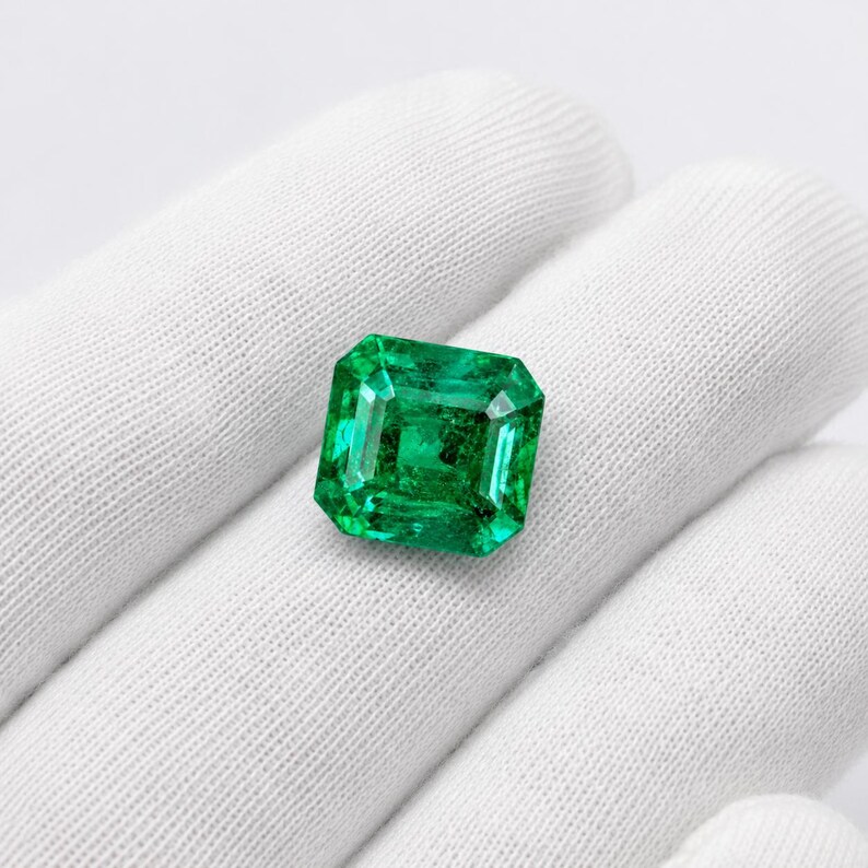 May include: A vibrant, emerald-green gemstone cut into a rectangular shape with faceted edges. The stone displays a deep, saturated green hue with internal clarity and light-reflecting surfaces, showcasing a classic step-cut design.