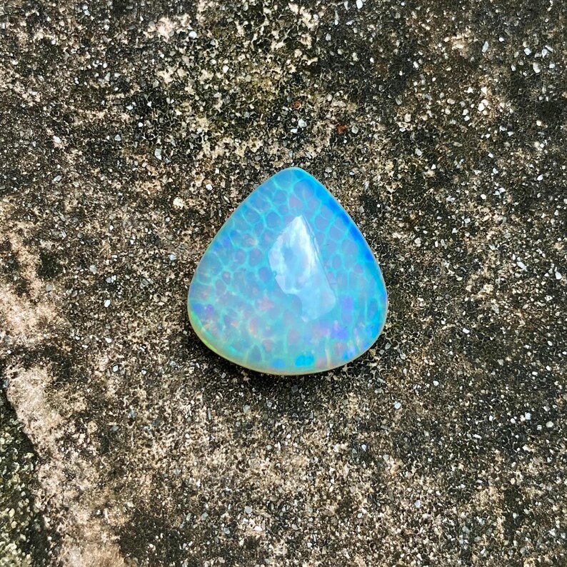 May include: A triangular, iridescent blue opal gemstone with a white center. The opal is sitting on a rough, gray surface.