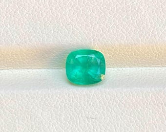 Puede incluir: Una gema de esmeralda verde vibrante con un corte de cojín. La gema está colocada sobre un fondo blanco.