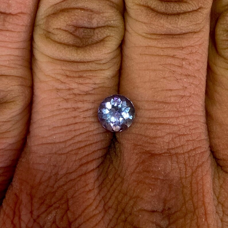 May include: A round, faceted, purple gemstone with a light blue hue.