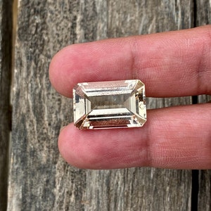 May include: A clear, rectangular, faceted gemstone with a light golden hue. The gemstone is held in a hand.