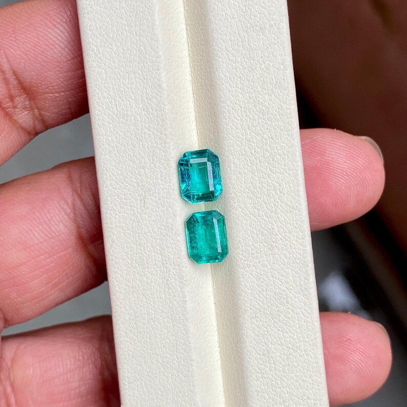 May include: Two rectangular emerald-cut gemstones with a vibrant green hue. The gems are displayed on a white surface, showcasing their clarity and brilliance. The stones are held by a person's hand, highlighting their size and color.