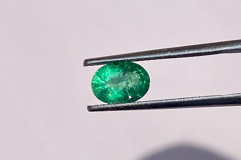 May include: A vibrant green emerald gemstone, held in tweezers, with a round oval shape.