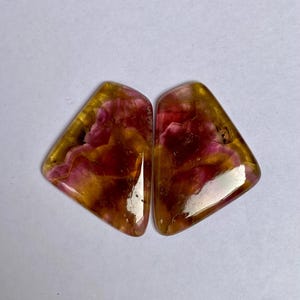 May include: Two polished gemstones with a triangular shape. The stones feature a blend of colours, including shades of pink, yellow, and brown. The gemstones have a glossy, reflective surface, and are set against a white background.