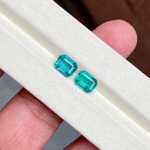 May include: Two rectangular, teal-colored gemstones with a bright, reflective surface. The gems are displayed on a white surface, showcasing their vibrant color and cut. The gems are held by a person's hand.