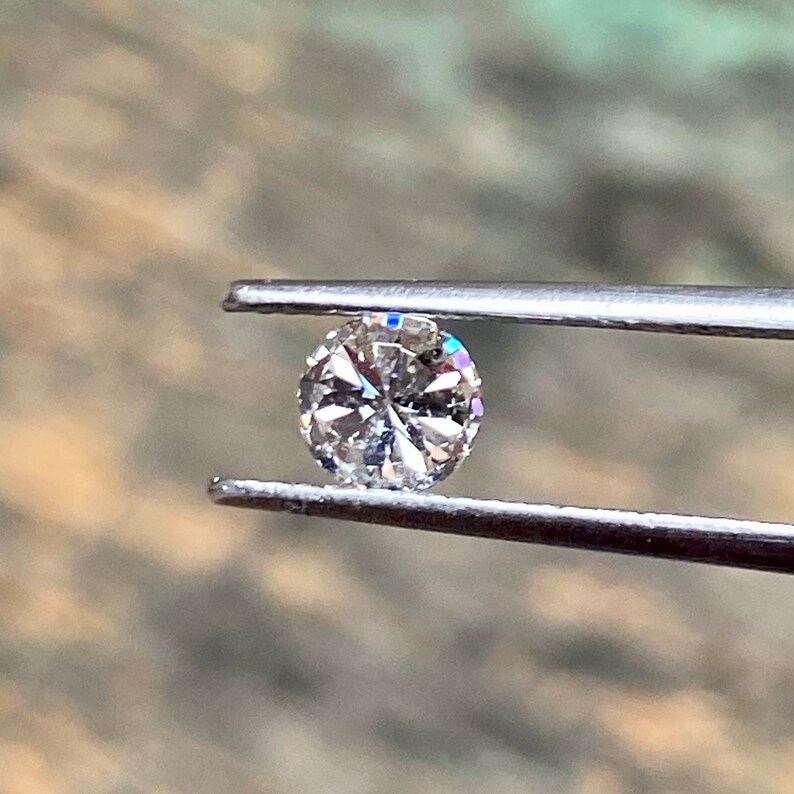 May include: A clear round diamond gemstone held by tweezers. The diamond has a faceted cut and is set in a silver setting.