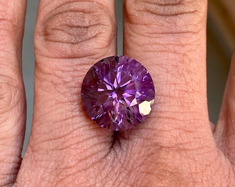 Könnte beinhalten: Ein großer, runder, violetter Amethyst mit Facettenschliff. Der Edelstein ist in einem Ring gefasst und wird an einem Finger getragen.