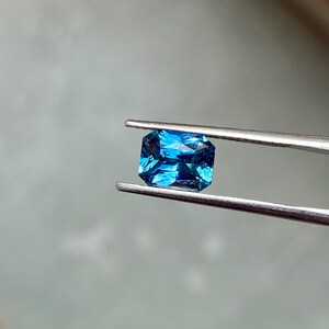 May include: A vibrant blue gemstone with a rectangular cut, held in tweezers. The gemstone has a deep, rich blue color and a slightly elongated shape.
