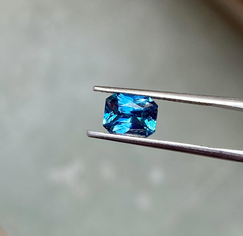 May include: A vibrant blue gemstone with a rectangular cut, held in tweezers. The gemstone has a deep, rich blue color and a slightly elongated shape.