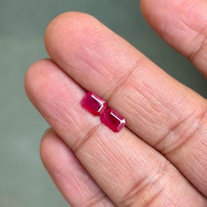 Könnte beinhalten: Zwei facettierte, rechteckige Rubine mit tiefroter Farbe. Die Edelsteine werden auf hellem Hintergrund präsentiert, um ihre Klarheit und Brillanz zu zeigen. Diese Rubine sind ideal für die Schmuckherstellung.