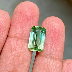 May include: A vibrant green gemstone with a rectangular emerald cut. The stone has a slight color gradient, transitioning from a lighter green to a darker green.