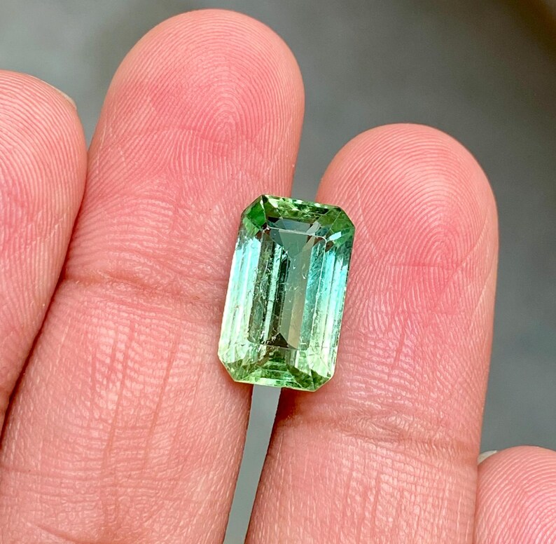 May include: A vibrant green gemstone with a rectangular emerald cut. The stone has a slight color gradient, transitioning from a lighter green to a darker green.