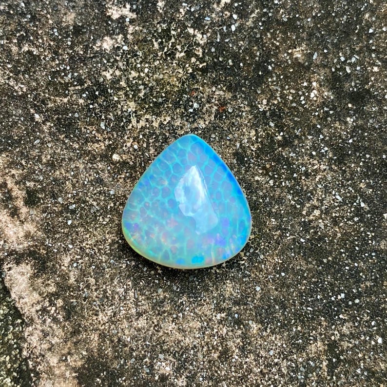 May include: A triangular iridescent opal gemstone with a blue and white color scheme. The opal is sitting on a rough, gray surface.