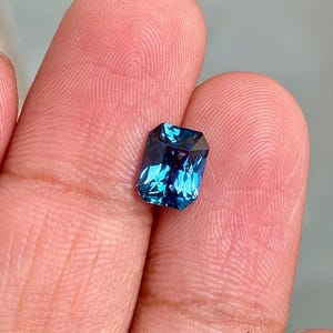 May include: A deep blue gemstone with a rectangular emerald cut. The gemstone is held in a hand.