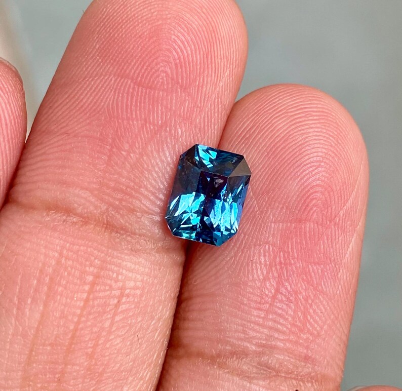 May include: A deep blue gemstone with a rectangular emerald cut. The gemstone is held in a hand.