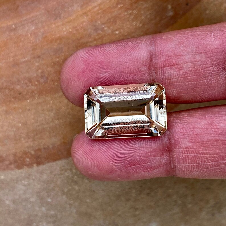 May include: A light brown, rectangular, faceted gemstone with a step cut. The gemstone is held in a hand.