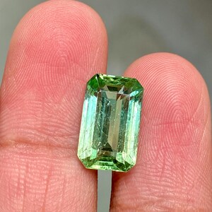 May include: A vibrant green emerald cut gemstone with a slight bluish hue. The gemstone is set in a clear, faceted setting.