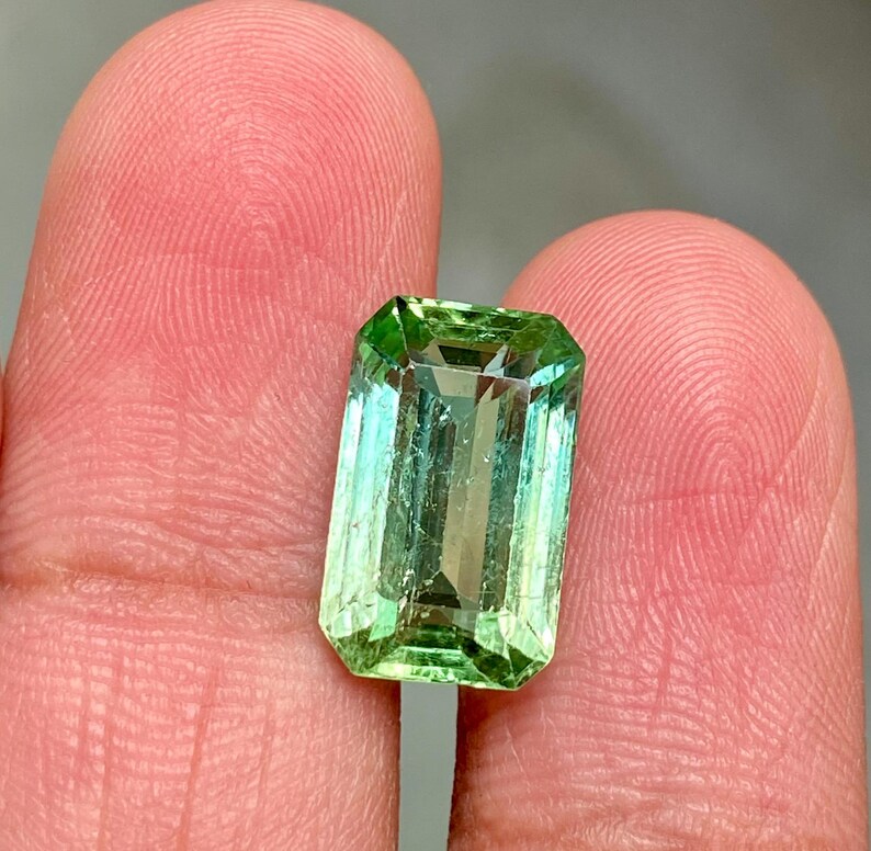 May include: A vibrant green emerald cut gemstone with a slight bluish hue. The gemstone is set in a clear, faceted setting.