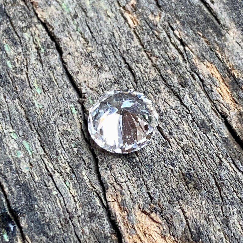 May include: A clear, round, faceted gemstone with a pointed top, resting on a piece of weathered wood.