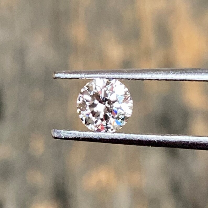 May include: A clear round diamond gemstone held in tweezers. The diamond is sparkling and reflecting light.