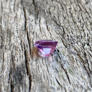 Puede incluir: Una piedra preciosa facetada de color p&uacute;rpura claro con forma cuadrada. La piedra preciosa est&aacute; sentada sobre un trozo de madera desgastada.