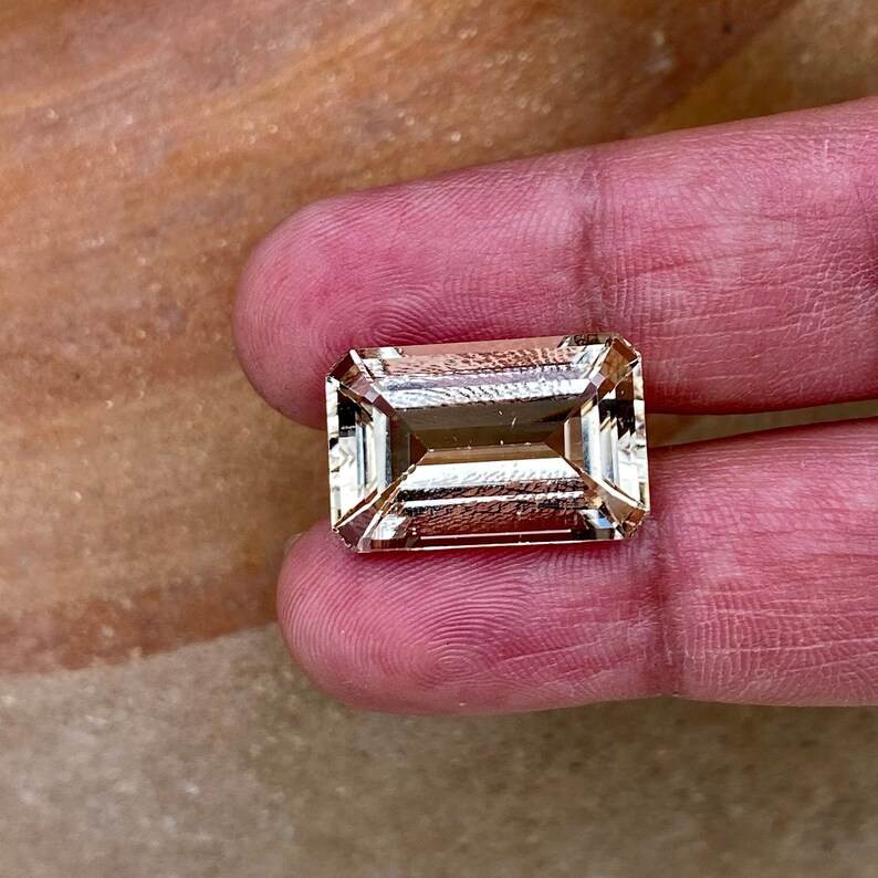 May include: A light brown, rectangular, faceted gemstone with a step cut. The gemstone is held in a hand.