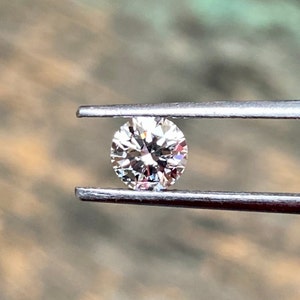 May include: A round, clear diamond gemstone held in tweezers. The diamond is faceted and reflects light.