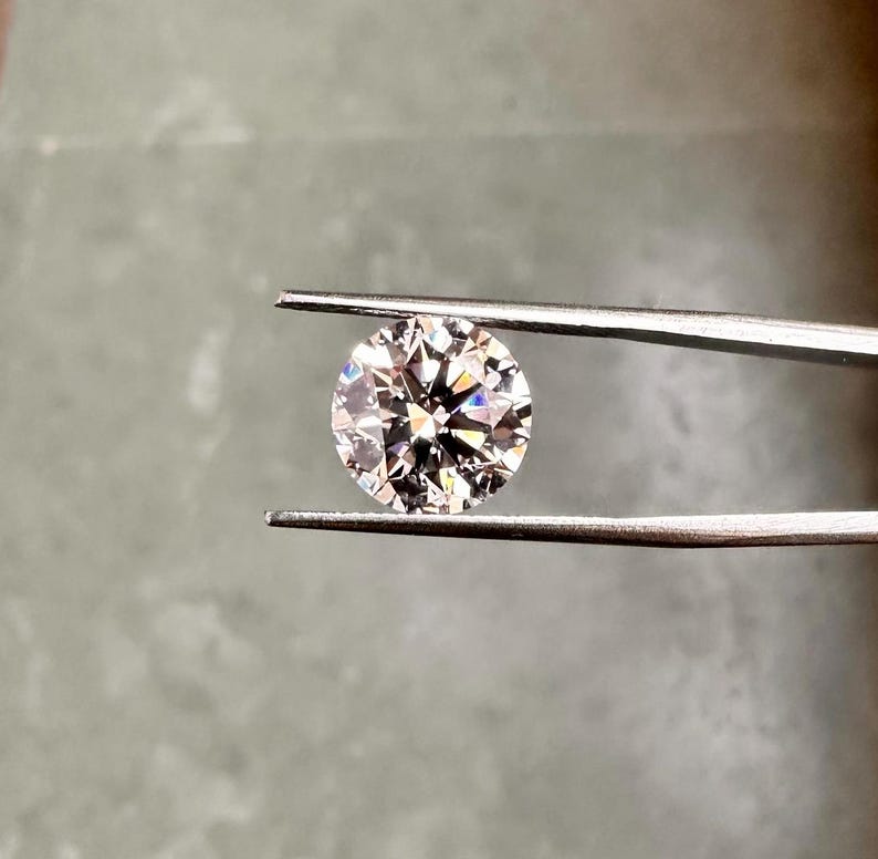 May include: A clear round diamond gemstone held by tweezers. The diamond has a brilliant cut and reflects light.