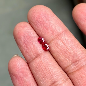 May include: Two small, oval-cut, bright red gemstones. The gems are displayed against a skin-toned background, showcasing their vibrant color and faceted surfaces. These gemstones could be rubies, perfect for jewelry making.