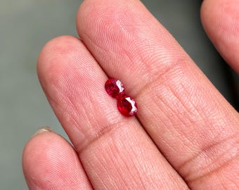 May include: Two small, oval-cut, bright red gemstones. The gems are displayed against a skin-toned background, showcasing their vibrant color and faceted surfaces. These gemstones could be rubies, perfect for jewelry making.
