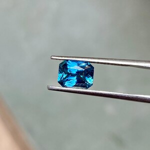 May include: A vibrant blue gemstone with a rectangular emerald cut, held by tweezers against a light gray background.