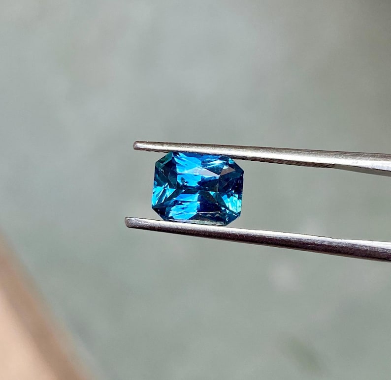 May include: A vibrant blue gemstone with a rectangular emerald cut, held by tweezers against a light gray background.