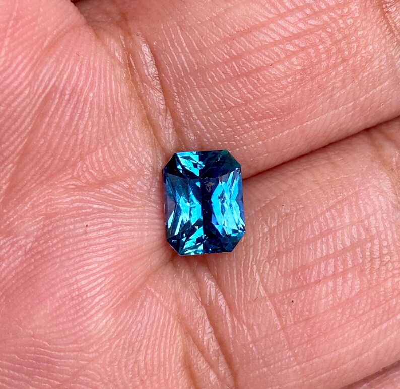 May include: A vibrant blue gemstone with a rectangular emerald cut. The gemstone is set in a bezel setting and has a high polish.