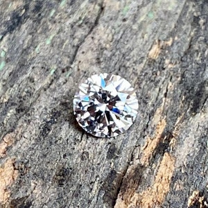 May include: A clear round diamond gemstone with many facets, set against a rough wooden background.