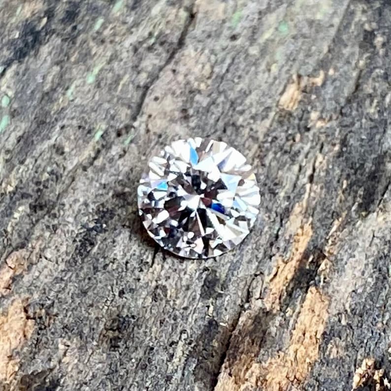 May include: A clear round diamond gemstone with many facets, set against a rough wooden background.