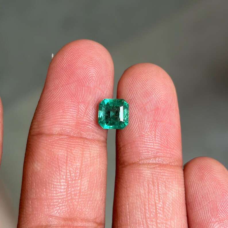 May include: A loose, square-cut emerald gemstone with a vibrant green hue and faceted surface. The stone displays internal clarity characteristics and light reflection across its polished top, showcasing a classic step-cut design with sharp corners.